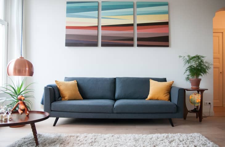 Blue sofa with yellow cushions, abstract triptych art, copper lamp, and plants in a modern living room.