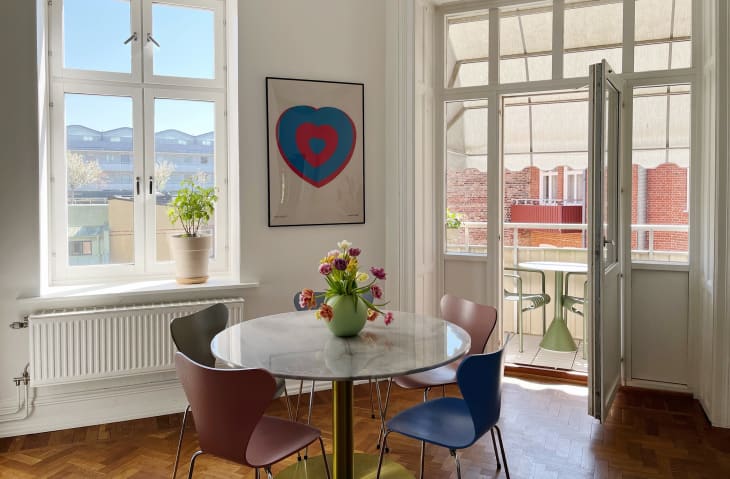 dining area with white walls, round marble dining table with different colored midcentury chairs, parquet wood floor, framed heart poster, window with potted plant on sill, doors open to balcony with table and chairs