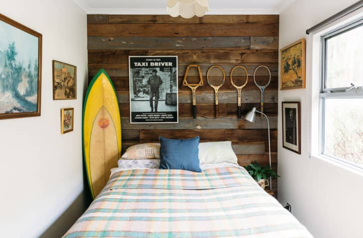 A surfboard, posters, and tennis rackets above a bed in a rustic bedroom