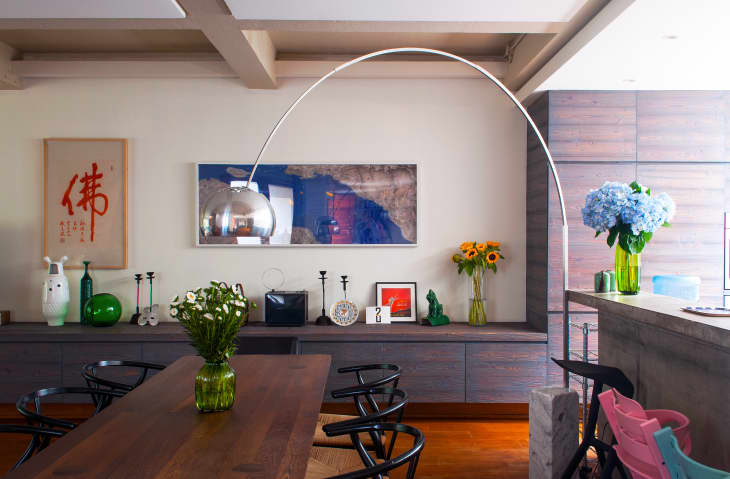 Dining room with wooden table, black chairs, green vase with flowers, art on walls, and large silver arc lamp.