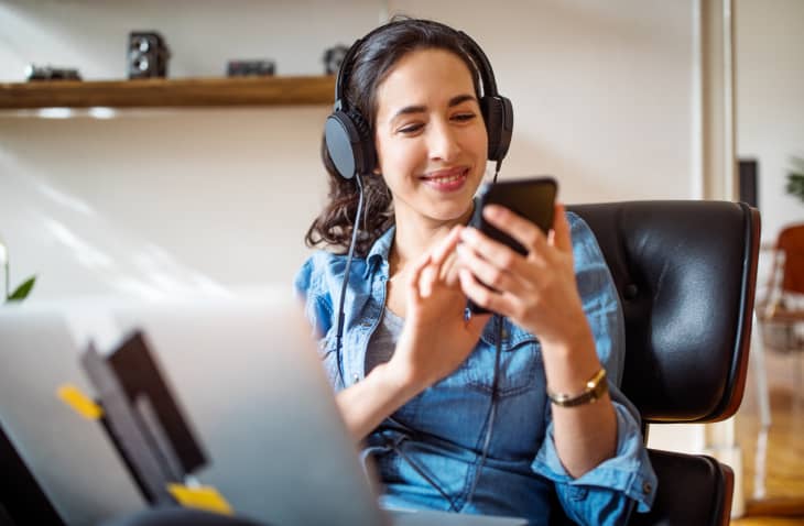 Happy young woman wearing headphones selecting a music playlist while working on laptop.