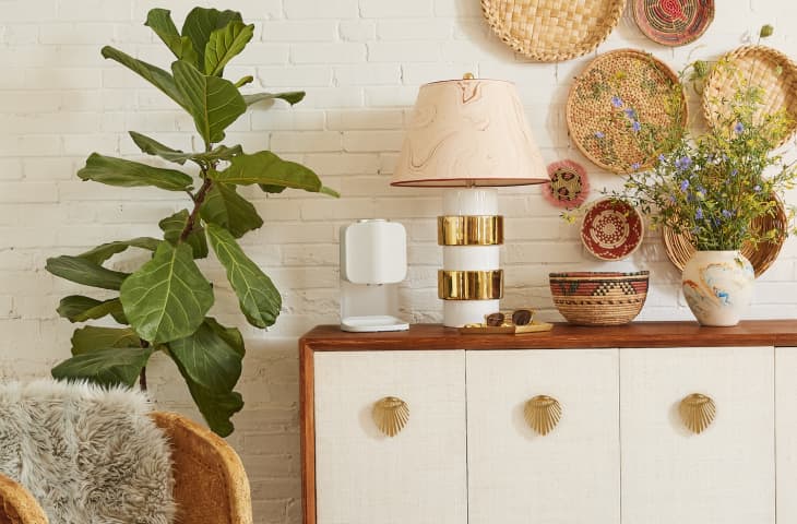 Sideboard with gold-accented lamp, woven baskets, vase with flowers, and a large leafy plant against a white brick wall.
