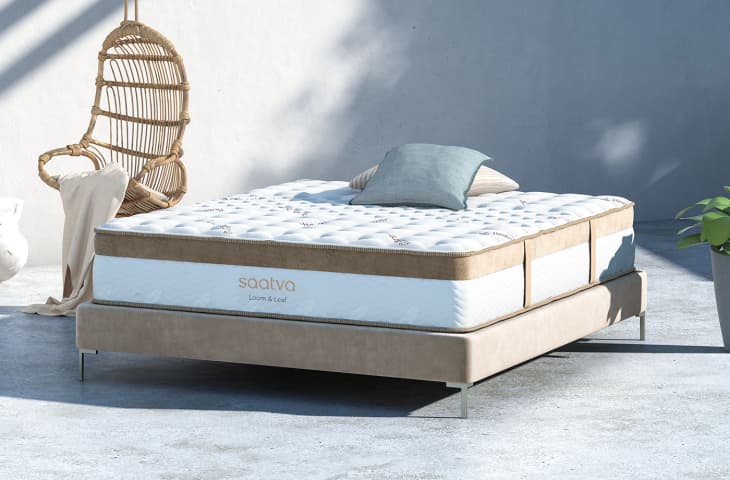 Saatva Loom & Leaf mattress on a beige bed frame, with a wicker chair and a plant nearby.