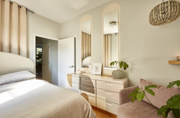 Cozy bedroom with beige curtains, a soft bed, two mirrors above a dresser, and a pink armchair with a potted plant.