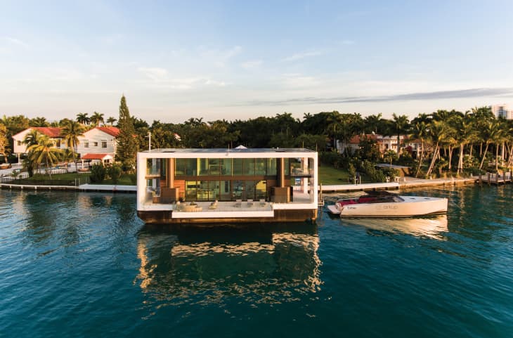 Modern floating house on water with large windows, docked boat, and lush greenery in the background.