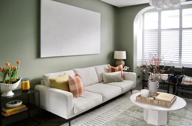 Living room with pale sage green walls, built in shelves, arched architectural details, modern art paintings, large window, pale gray sofa, natural textile rug, white "balloons" style light fixture