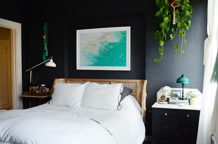 Bedroom with dark walls, white bedding, a rattan headboard, hanging plant, and a turquoise beach painting above the bed.