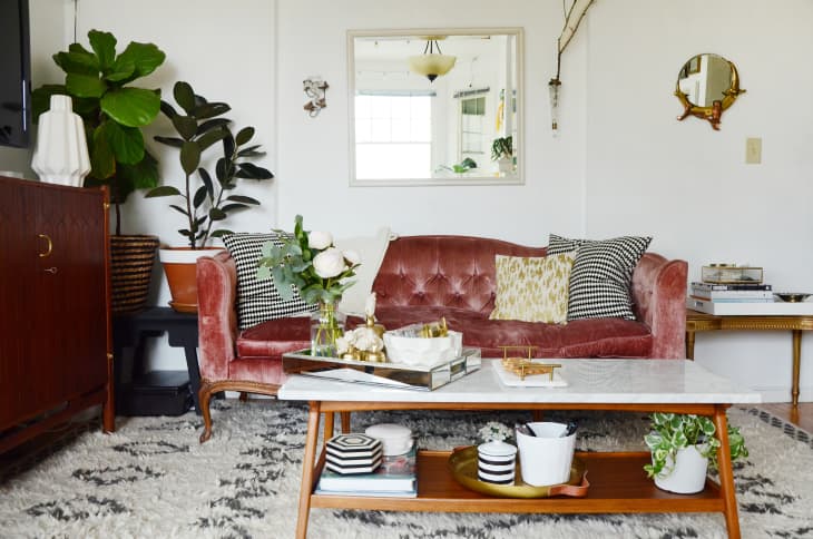 Velvet pink sofa with patterned cushions, marble coffee table, plants, and decorative mirrors in a cozy living room.