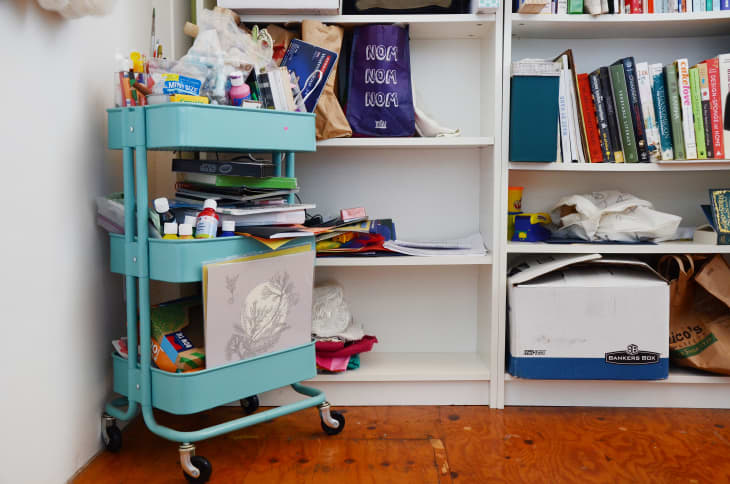 Turquoise rolling cart with art supplies next to a white bookshelf filled with books and storage boxes.