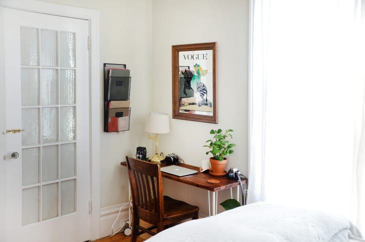 Small home office with wooden desk, chair, potted plant, lamp, and Vogue poster on wall.