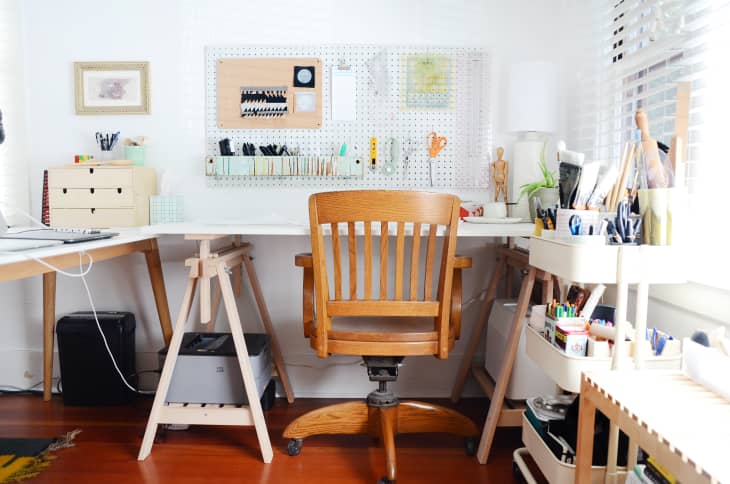 Home office with wooden chair, pegboard with tools, desk with storage boxes, and art supplies on shelves.