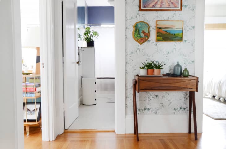 Hallway with wooden console table, potted plants, and framed art on floral wallpaper, leading to a bright room.