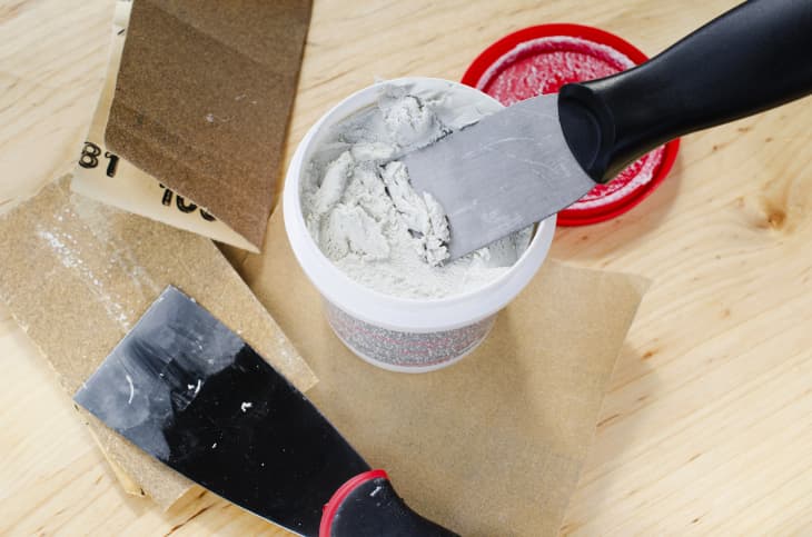 spackle knife in white spackle tub on wood surface.another knife and sandpaper in photo