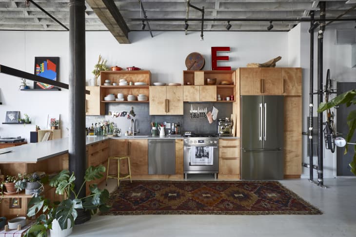 Industrial-style kitchen with wooden cabinets, open shelving, stainless steel appliances, and a patterned rug.