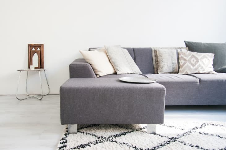 Gray sectional sofa with assorted pillows, small side table, and decorative wooden panel in a minimalist living room.