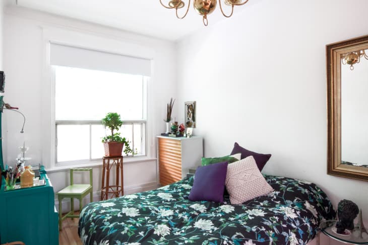 Bedroom with floral bedspread, purple and white pillows, green dresser, potted plant, and large mirror.