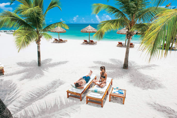 Two people relaxing on lounge chairs under palm trees on a sandy beach with turquoise ocean in the background.