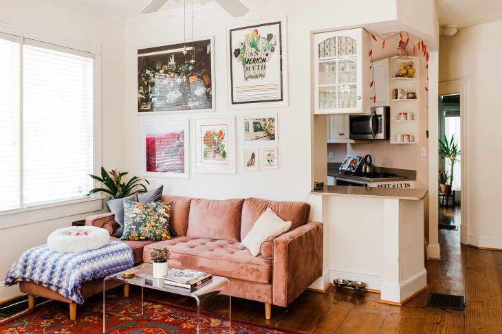 Cozy living room with a pink sofa, colorful pillows, wall art, and a small kitchen nook with open shelving.