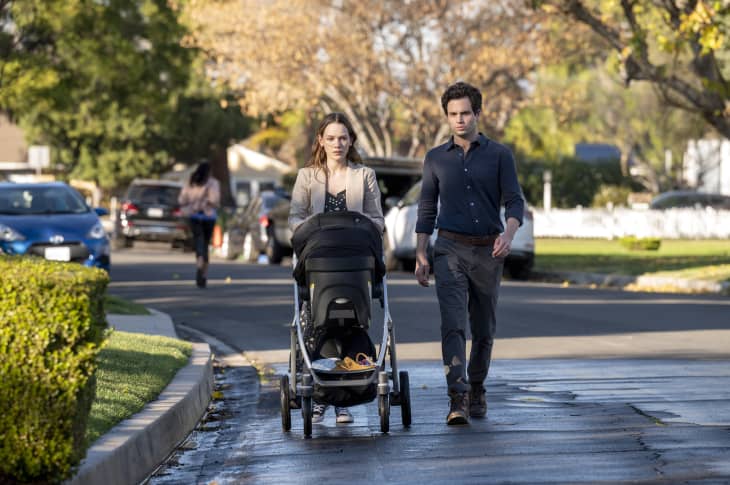 A man and woman walking on a suburban street with a baby stroller, surrounded by trees and parked cars.