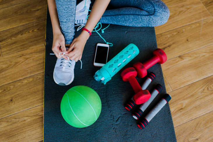 Person tying shoelaces on exercise mat with dumbbells, green ball, water bottle, and smartphone nearby.