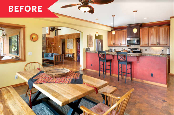 Open-plan kitchen and dining area with wooden table, red accent wall, and bar stools, leading to a cozy living room.