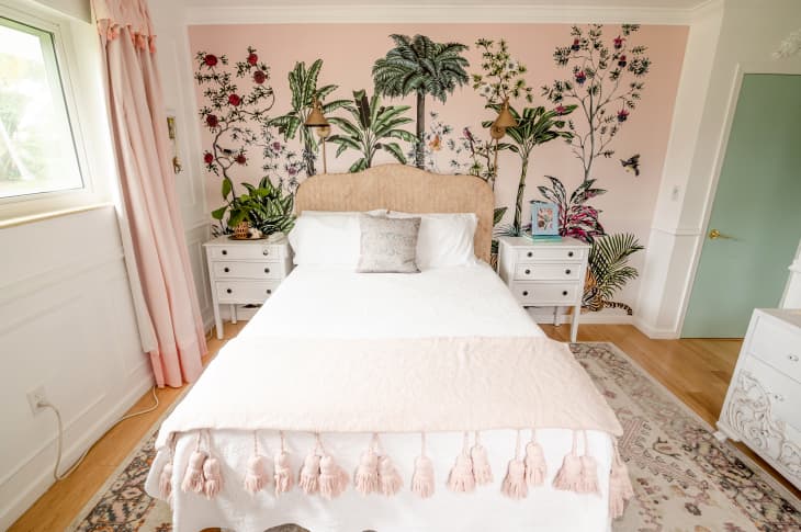 blush pink, white, and green bedroom with pink wallpaper that has trees and plants on it. 2 white bedstands with drawers flank the bed, which has a white bedspread and light pink throw blanket. Tall pink curtains frame a large window