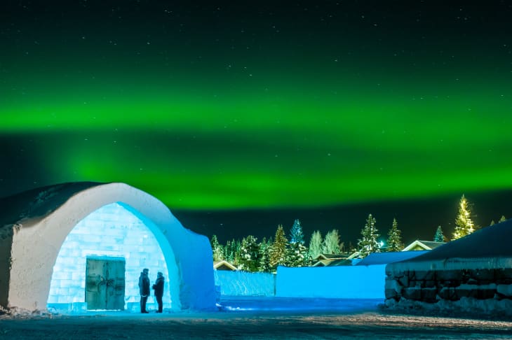 Two people stand outside an illuminated igloo under the green aurora borealis.