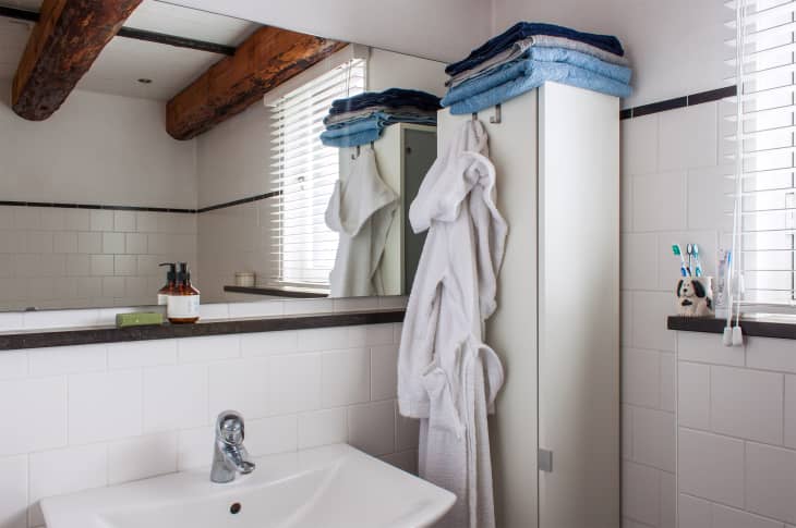 Bathroom with wooden beams, white tiles, a sink, soap dispenser, towels, bathrobe, and toothbrushes by the window.