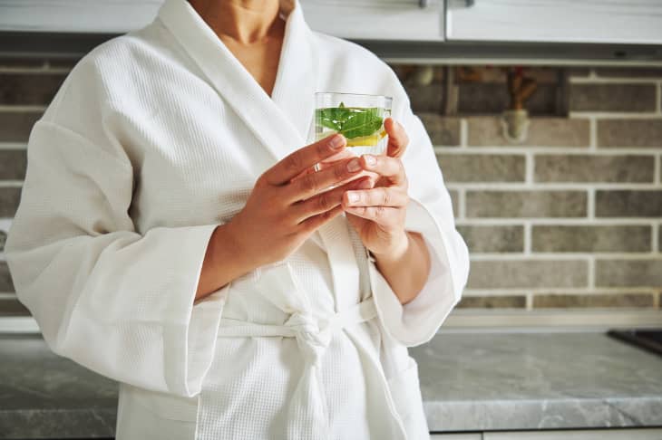 Cropped view of unknown woman in white waffle bathrobe holding a glass with lemon and mint water. Concepts of healthy lifestyle and diet.