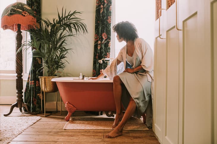 Woman perches on the side of an elegant red roll top bathtub, and waits for it to fill up. She wears a silky robe. Light floods through the window, backlighting the relaxing scene and giving it a dreamy vibe.