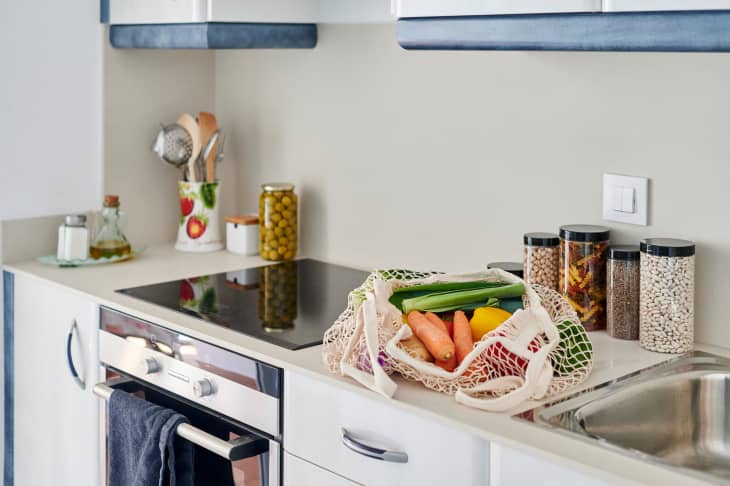Modern kitchen with induction stove, utensils, jars of spices, and a mesh bag of fresh vegetables on the counter.