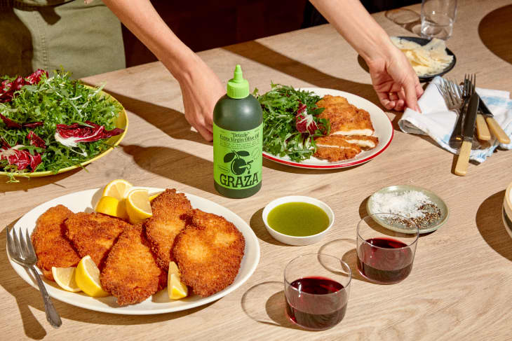 Breaded cutlets with lemon wedges, green salad, olive oil bottle, and two glasses of red wine on a wooden table.