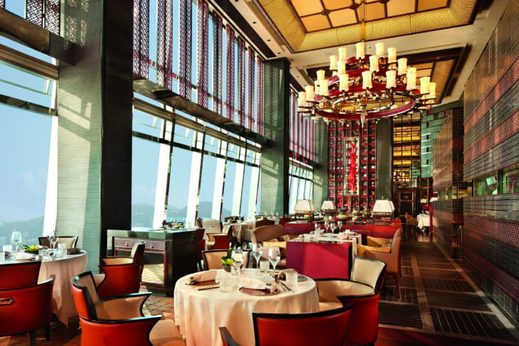 Elegant restaurant with round tables, red chairs, large chandelier, and floor-to-ceiling windows offering a scenic view.