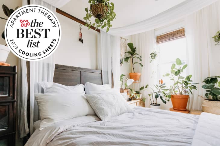 Bed with pale gray and white cotton bedding, wood headboard, white walls, sheer curtains behind bed, window letting in lots of light, multiple plants. Seal in upper left corner reads "Apartment Therapy the Best List 2023 Cooling Sheets"
