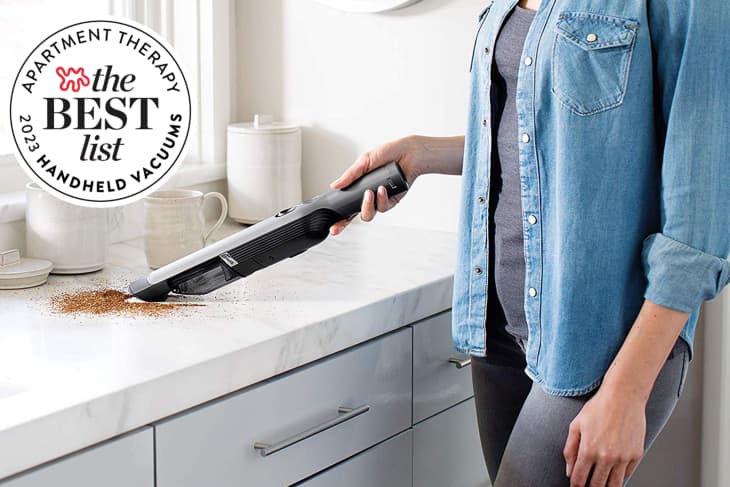 Woman using Shark handheld vacuum to vacuum up coffee grounds on counter. Seal in upper left corner says "Apartment Therapy The Best List 2023 Handheld Vacuums"
