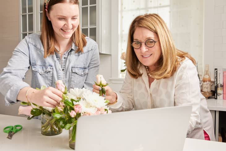 Two women arranging flowers in a kitchen, smiling and looking at a laptop.