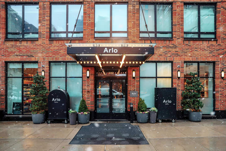 Entrance of Arlo Hotel with brick facade, large windows, potted plants, and snow falling.