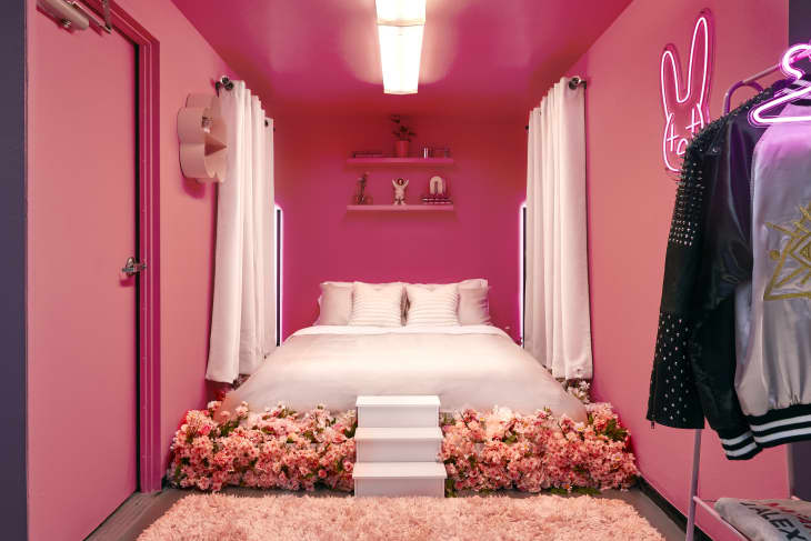 Pink bedroom with white bed, floral decor, neon bunny sign, and studded jackets on a rack.