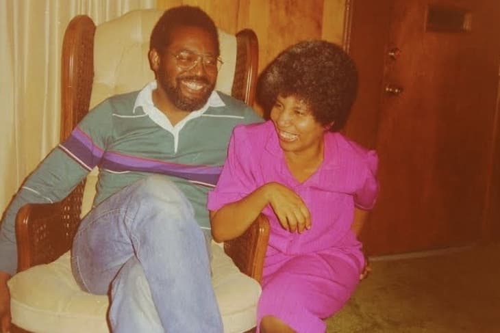 Man in striped sweater and woman in pink dress sitting on a cushioned chair, smiling and laughing together.