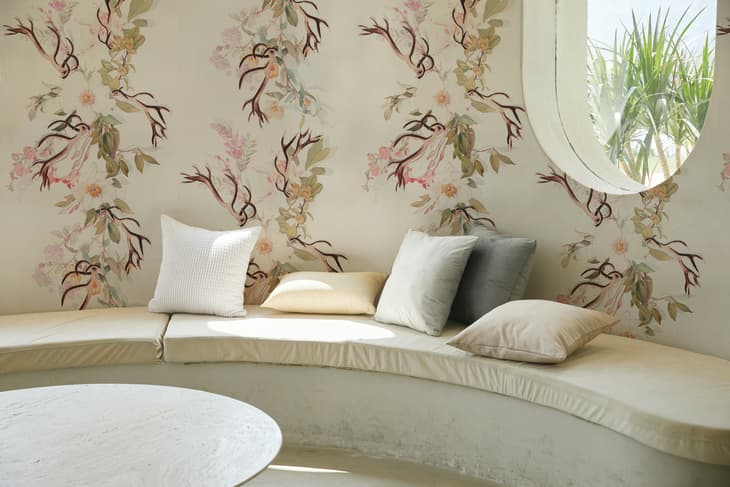 Curved bench with pastel cushions against floral wallpaper and round window.