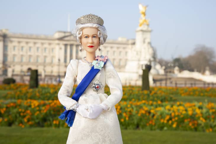 A doll in a white gown with a blue sash stands in front of Buckingham Palace, surrounded by orange flowers.
