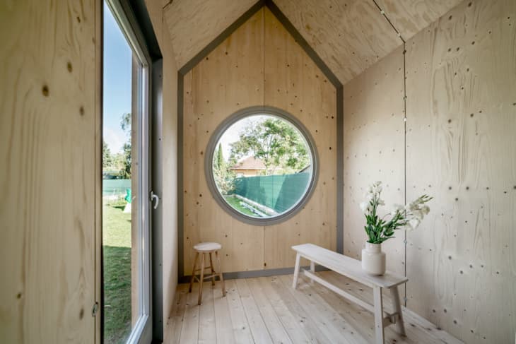 Minimalist wooden room with round window, bench, stool, and vase of white flowers.
