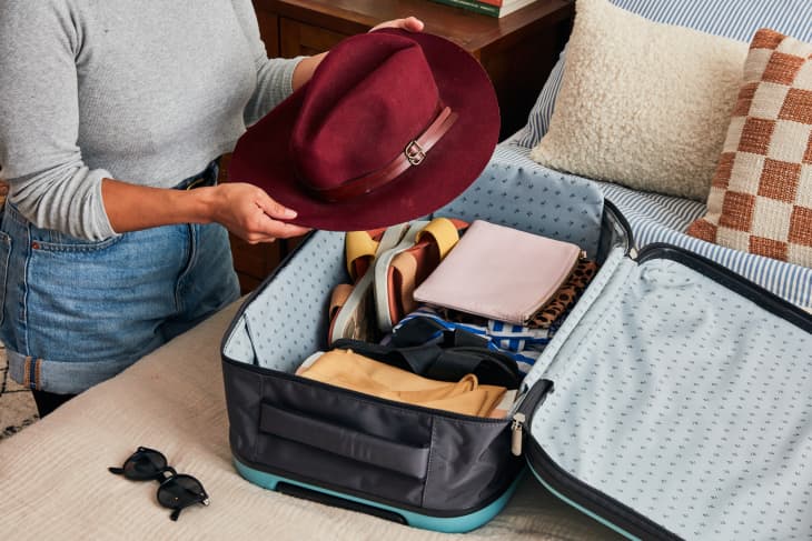 semi closeup shot of a person holding a red fedora hat over a grey suitcase thats on a bed with beige sheets and sunglasses laying next to it