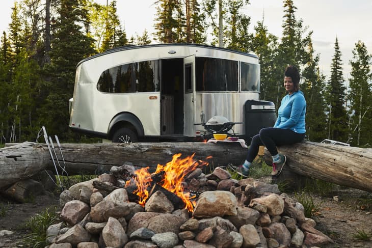 Woman sitting on a log by a campfire with a silver camper in a forest setting.
