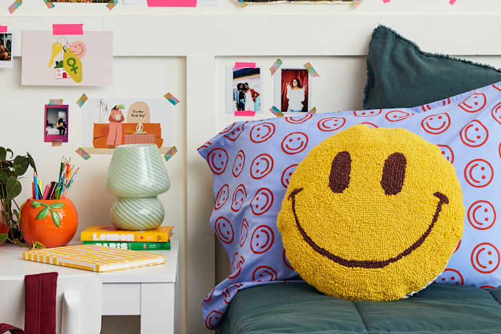 semi wide shot of a bed with teal sheets, blue pillowcase with red smiley faces on it, a smiley face pillow and a white desk with a red backpack hanging off white chair, a green lamp and supplies on top of desk, and a wall with different photos and art