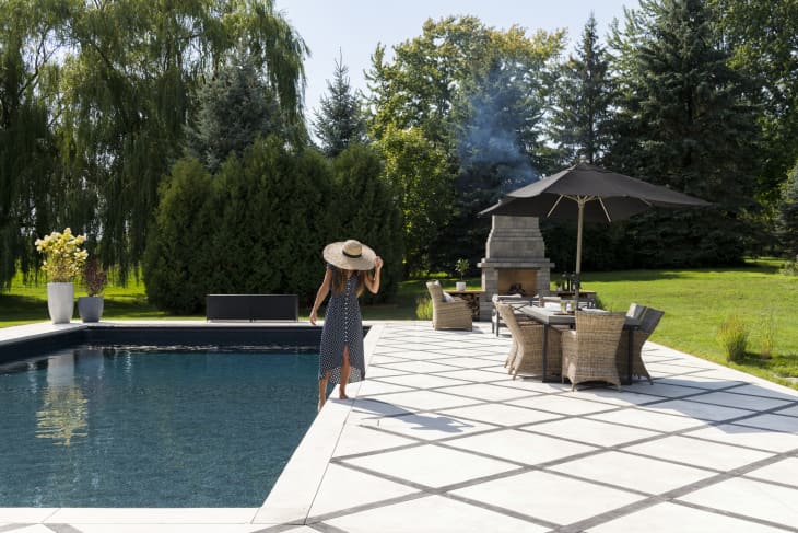 Woman in a sun hat walking by a pool, with patio furniture and a fireplace under a black umbrella in a garden setting.
