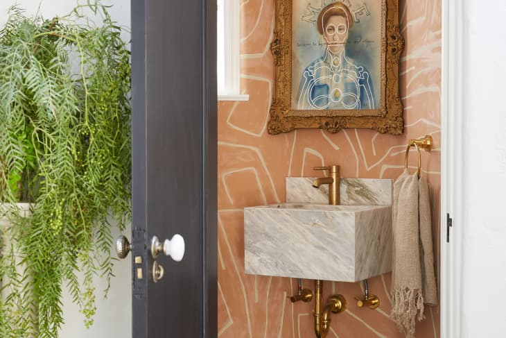 A powder room with orange wallpaper that has white squiggly lines on it, and a marble sink below a framed portrait. The portrait has a gold frame, and the sink has gold hardware. There's a beige towel with a gold towel ring beside it.