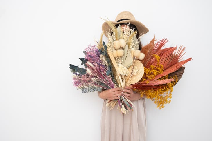 Person in a beige dress and hat holding a large bouquet of colorful dried flowers.