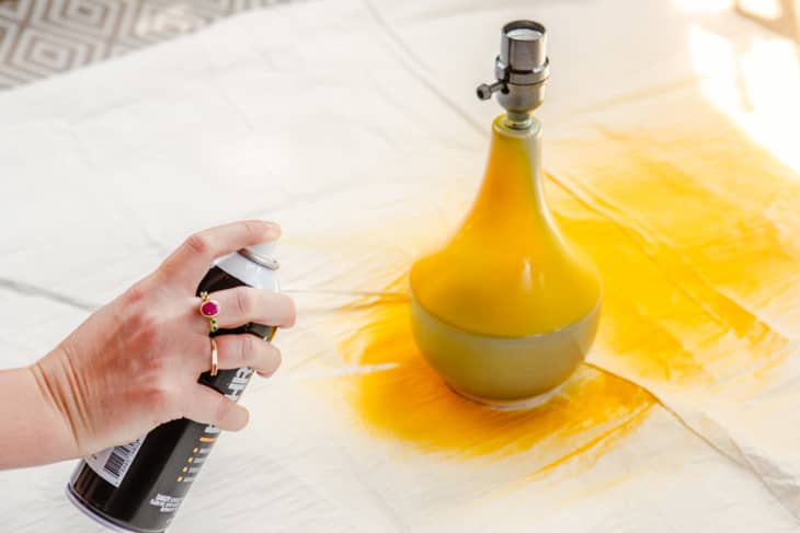 Person spraying yellow spray paint sitting on a small table lamp. Drop cloth underneath