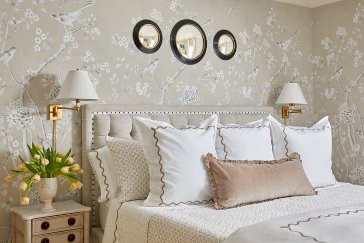 Elegant bedroom with floral wallpaper, beige headboard, white pillows, and gold wall lamps.